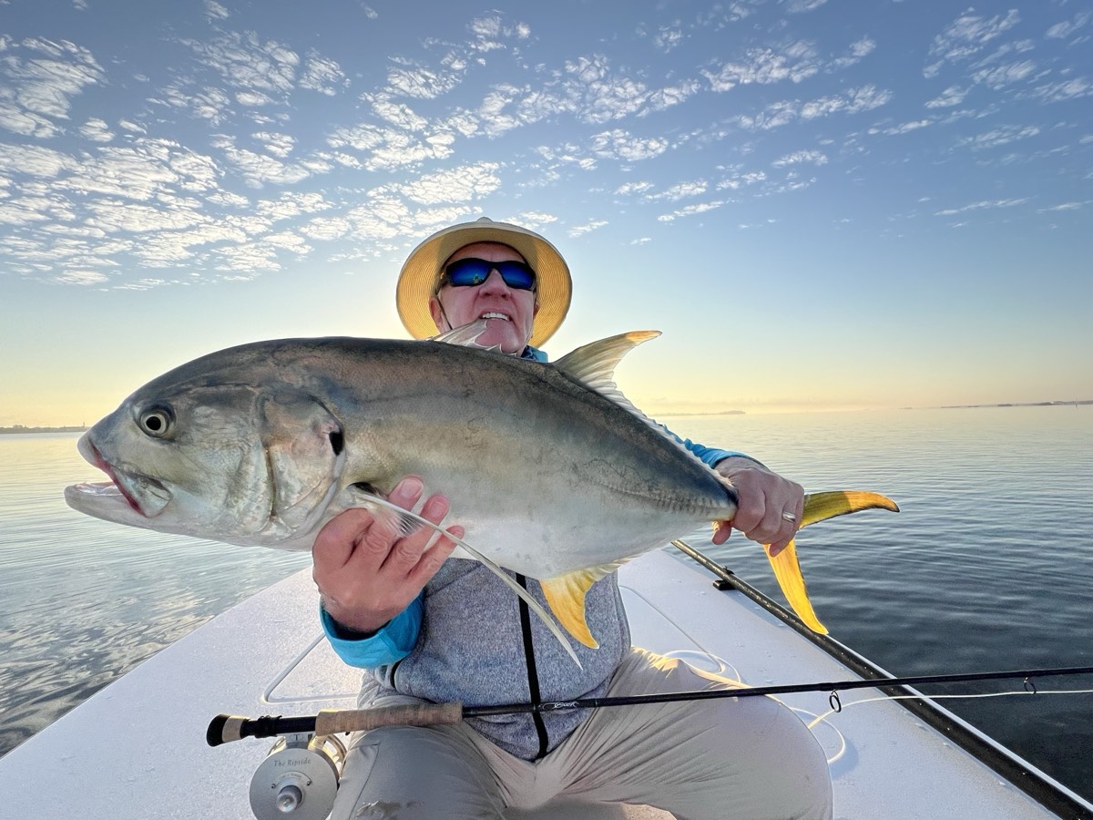 Brian Pitre seated on a boat, holding a large silver fish with a yellow tail, under a bright scattered sky.