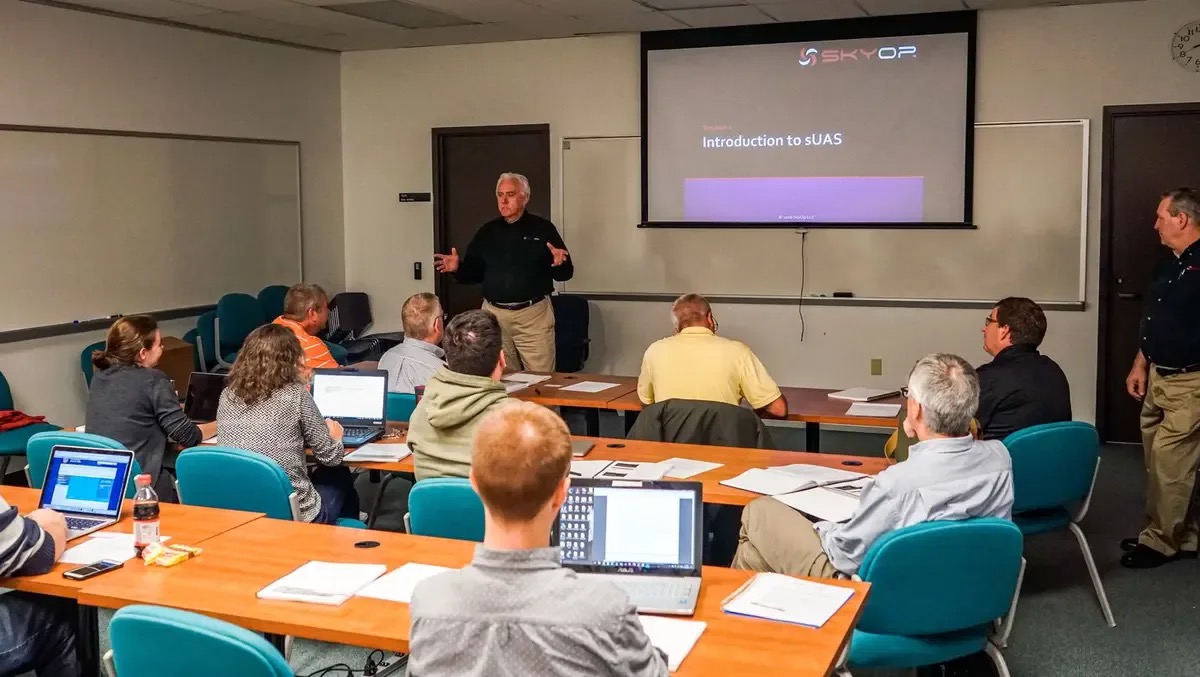 Brian Pitre standing at the front of a classroom, teaching a SkyOp Introduction to sUAS session. A projector behind him shows the SkyOp name.
